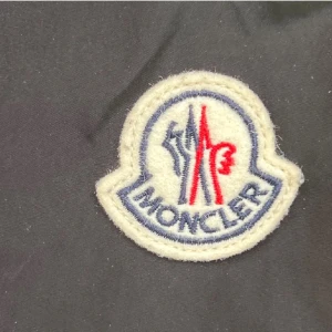  Moncler windbraker  - Marinblå vindjacka från Moncler med huva och dragkedja framtill. Jackan har Moncler-logga på ärmen och snygga detaljer med snörning i huvan. Materialet är lätt och känns sportigt, perfekt för blåsiga dagar. Klassisk och stilren design med diskreta färgdetaljer i huvan.