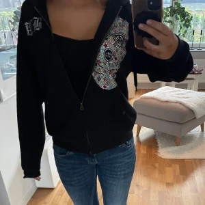 Bvaldi zip up hoodie svart - Svart hoodie med dragkedja och huva. På framsidan finns ett stort motiv av en mask i färgglada strasstenar och text i strass på bröstet. På ryggen står det 'BANK ROBBER' i strass. Hoodien har normal passform och långa ärmar. Perfekt statement-plagg för dig som gillar unika detaljer.