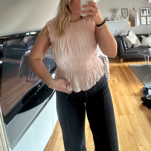 Ljusrosa blus med smock och volang - Supersöt ljusrosa blus med smockad midja och volangkant nertill. Ärmlös modell med rund halsringning och diskret dragkedja i sidan. Tunt, lätt tyg som ger en luftig känsla. Lite slitage på insidan men inget som syns när man har tröjan på sig