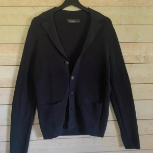 Mörkblå stickad kofta från Dressmann - Säljer en mörkblå stickad kofta från Dressmann i storlek S. Koftan har klassisk krage, knäppning med fyra knappar och två praktiska fickor framtill. Perfekt för lager-på-lager och chill höst- eller vinterstil.