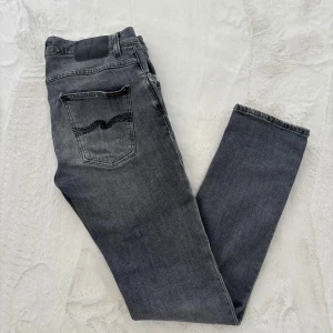 Nudie Jeans Grim Tim grå jeans - Säljer ett par Nudie Jeans Grim Tim i blekgrå tvätt. Klassisk femficksmodell med svarta sömmar på bakfickan. Jeansen är raka i modellen och tillverkade i 98% bomull och 2% elastan för lite stretch. Perfekta för dig som gillar en avslappnad men snygg look. Lagningar behövs.