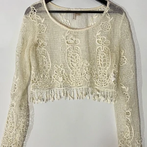 Cremvit spetsig croppad blus med fransar - Superfin cremvit croppad blus med spets och broderade blommor. Långärmad modell med transparent känsla och snygga fransar längs nederkanten. Perfekt för dig som vill sticka ut med en romantisk och bohemisk vibe.