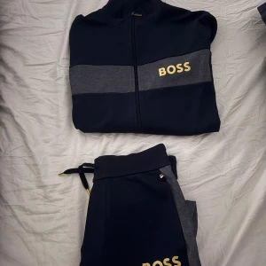 Mörkblå ziptröja och mjukisbyxor från BOSS - Snygg mörkblå ziptröja och mjukisbyxor  från BOSS med grå panel över bröstet och guldig BOSS-logga. Tröjan har hög krage, dragkedja framtill och är perfekt för en sportig look. Materialet känns mjukt och stretchigt, troligen polyesterblandning.