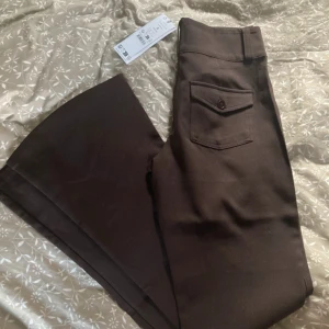 Bruna bootcut byxor med fickor - Snygga bruna bootcut-byxor från Gina tricot med låg midja och två bakfickor med lock och knapp. Byxorna är bootcut och stängs med två knappar framtill. Perfekta för dig som vill ha en trendig och stilren look.