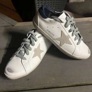 Golden Goose Superstar vita sneakers - Golden Goose Superstar sneakers i vitt skinn med grå mockastjärna på sidan, grå snörning och mörkblå häl. Klassisk låg modell med rund tå och platt sula. Snygg kontrast mellan material och färger, perfekt för en trendig streetstyle.