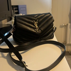 Svart quiltad axelväska från YSL - Säljer en svart quiltad axelväska från YSL med ikoniskt guldigt logospänne framtill. Väskan är i skinn och har justerbar axelrem samt flera fack inuti, inklusive ett med dragkedja. Perfekt för dig som vill ha en stilren och lyxig accessoar.