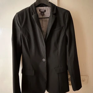 Ny Svart kavaj från Lindex, stl 36 - Klassisk svart kavaj från Lindex i storlek 36 Enkelknäppt modell med två fickor fram och en bröstficka. Snygg passform med figursydd siluett och helfodrad insida. Perfekt att styla med skjorta eller t-shirt för en clean look.