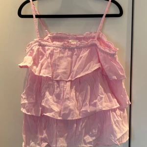 Rosa volangtop från Gina Tricot - Supersöt ljusrosa linnetopp från Gina Tricot med tunna axelband och flera lager volanger. Toppen har en luftig och flowy känsla, perfekt för varma dagar. Tillverkad i bomull för en skön och lätt känsla.