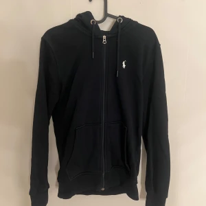 Svart hoodie från Ralph Lauren - Svart hoodie från Ralph Lauren med dragkedja och huva. Klassisk design med vit logga på bröstet och praktiska fickor framtill. 