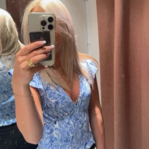 Blommig blå omlottopp - Supersöt topp med blått blommönster och omlottdesign. Djup v-ringning och korta ärmar ger en fräsch vibe. Perfekt för dig som gillar romantiska detaljer och vill sticka ut lite extra.