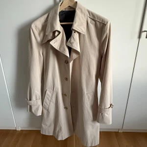 Beige trenchcoat med bälte - Stilren beige trenchcoat från SAND. Perfekt för vår och höst när du vill ha en clean look. Endast använd 2-3 gånger. Ordinarie pris 3 499 kr. Längd: 98 cm, ärm: 65 cm, bröst: 60 cm, midja 57 cm. Jag är 186 cm, 86 kg