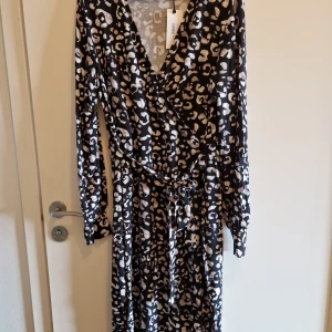 Leopardmönstrad omlottklänning Indiska - Snygg långärmad omlottklänning från Indiska med leopardmönster i svart, beige och vitt. Klänningen har knyt i midjan och v-ringning, vilket ger en trendig och bekväm passform. Perfekt för dig som gillar djurmönster och vill sticka ut.