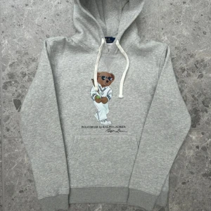 Grå hoodie Polo Bear Ralph Lauren - Är i M men är liten i storleken så den passar S. Säljer en grå hoodie från Polo Ralph Lauren med ikoniska Polo Bear-trycket på bröstet. Tröjan har huva med vita snören och en stor magficka. Perfekt för dig som gillar streetstyle och vill ha något bekvämt och snyggt.