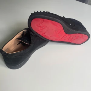 Christian Louboutin svarta sneakers med nitar - Svarta sneakers i mocka från Christian Louboutin med ikonisk röd sula och svarta nitar på tån.  Med några defekter som visas på bilderna, skorna är använda i ungefär 1 år, fast än dess skador så kan de användas och fortfarande ge dig en lyxig och edgy look! Skriv gärna om du har frågor