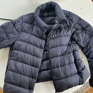 Svart dunjacka från Uniqlo XS - Säljer en svart, ultralätt dunjacka från Uniqlo i storlek XS. Jackan har en klassisk pufferdesign med horisontella sömmar, hög krage och dragkedja framtill. Perfekt för kyliga dagar och enkel att bära med sig tack vare sin lätta vikt.originella priset är runt 800/900kr lite små fjädrar som man kan se på bilden som sticker ut fast inget märkvärdigt 