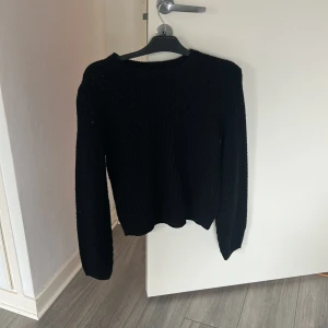 Svart stickad tröja med struktur - En svart stickad tröja med snygg struktur och rund halsringning. Tröjan har långa ärmar och ett diskret mönster i stickningen som ger en cool och stilren look. Perfekt att slänga på sig när det är kyligt ute.