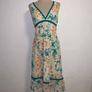 Blommig sommarklänning från Åhléns - Supersöt blommig klänning från Åhléns med v-ringning. Klänningen har ett färgglatt fjärilsmönster i turkos, rosa och gul på ljus botten, samt dekorativa band i turkos. Perfekt för varma dagar.