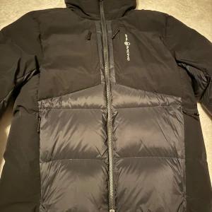 Svart dunjacka från Sail Racing - Säljer en svart dunjacka från Sail Racing med 600 FP dunfyllning. Jackan har huva, dragkedja framtill och flera praktiska fickor med dragkedja. Perfekt för kalla dagar och riktigt snygg streetstil. Logga på bröstet och insidan.