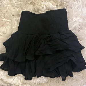 Svart volangkjol - Säljer en svart kortkjol med flera lager volanger. Kjolen har en flowy och luftig känsla, perfekt för att skapa en cool och trendig look. Materialet är lätt och ger snygg rörelse när du går.