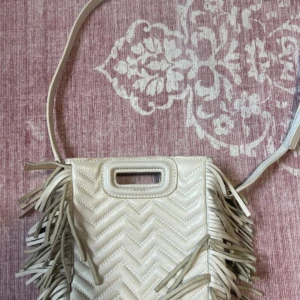 Vit axelväska med fransar från Maje - Supersnygg vit axelväska från Maje i skinn med quiltad zickzack-design och långa fransar på sidorna. Väskan har både axelrem och ett coolt handtag upptill. Perfekt för dig som vill sticka ut med en unik accessoar.