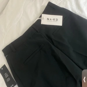 Svarta kostymbyxor från NA-KD - Stilrena svarta kostymbyxor från NA-KD med hög midja och raka ben. Byxorna har pressveck, hällor för bälte och diskreta fickor bak. Perfekta för en clean och snygg look. Materialet är mjukt och följsamt, vilket gör dem bekväma hela dagen.