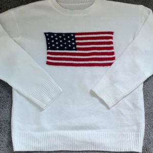 Vit stickad tröja med USA-flagga - Snygg vit stickad tröja med lång ärm och ribbade muddar. På bröstet finns en stor USA-flagga i blått, rött och vitt som ger en cool och unik look. Perfekt för dig som gillar statement-plagg och vill sticka ut lite extra.