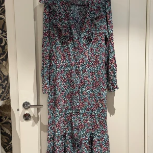 Blommig långklänning från By Malina - Säljer en färgglad långklänning från By Malina med blommigt mönster i blått, rosa, vitt och orange. Klänningen har volangdetaljer vid halsen och nederkanten, v-ringning och långa ärmar. Perfekt för dig som gillar boho-vibe och vill sticka ut med en unik look.