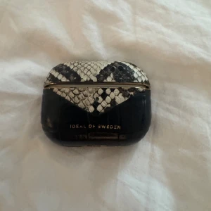 Svart och vit AirPods-fodral Ideal of Sweden - Snyggt AirPods-fodral från Ideal of Sweden i svart och vitt med ormmönster och gulddetaljer. Fodralet har en lyxig känsla med reptilstruktur och passar perfekt för att ge dina hörlurar en trendig look.