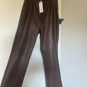 Bruna skinnbyxor från NLY Trend - Snygga bruna skinnbyxor från NLY Trend med rak passform och slits nertill på benen. Byxorna har hög midja och dragkedja i sidan för enkel påtagning. Materialet är mjukt.