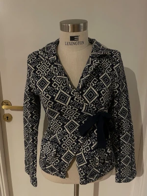 Mönstrad omlottkofta från Lely Wood - Snygg omlottkofta från lely wood i mörkblått och vitt med geometriskt mönster. Koftan har lång ärm, knytband i midjan och är stickad i mjukt material. Perfekt för dig som gillar detaljer och vill ha en unik look.