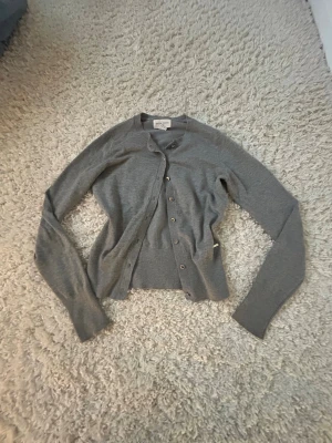 Grå kofta från Holly & Whyte XS - Snygg grå kofta från Holly & Whyte by Lindex i storlek XS. Koftan har rund hals, långa ärmar och knappar framtill. Perfekt att slänga över en t-shirt eller linne för en chill look. Materialet är mjukt och känns skönt mot huden.