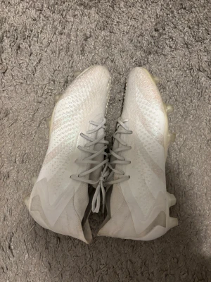 Vita fotbollsskor från Adidas - Säljer ett par vita adidas fotbollsskor med snörning och låg profil. Skorna har en rund tå och platt sula med dobbar för gräsplan. Ovandelen är i syntetmaterial med diskreta mönster och adidas-logga på hälen. Perfekt för dig som vill ha stilrena och lätta skor på planen. Tveka inte att höra av er vid några frågor eller funderingar🤗