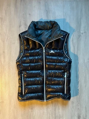 Mörkblå dunväst från J.Lindeberg - Snygg mörkblå dunväst från J.Lindeberg med hög krage och silvriga dragkedjor. Västen har quiltad design, två fickor med dragkedja och diskret logga på bröstet och nacken. Perfekt att slänga över en hoodie för en cool look. Den ser svart ut på bilderna men den är mörkblå. Det står ingen storlek men jag skulle säga S till M