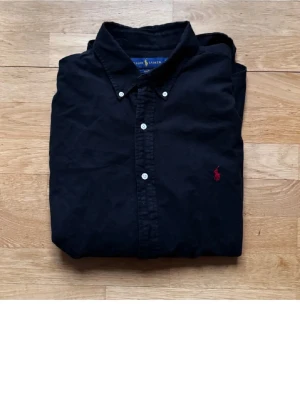 Svart skjorta från Ralph Lauren M/L - Svart skjorta från Ralph Lauren i slim fit med klassisk krage och vita knappar. Broderad röd logga på bröstet ger en snygg kontrast. Skjortan är långärmad och tillverkad i mjuk bomull som känns skön mot huden.