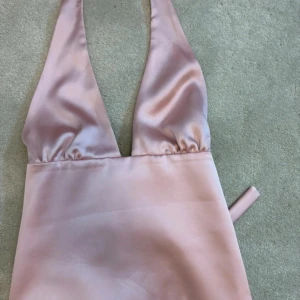 Rosa satin topp med djup v-ringning - Supersnygg rosa topp i satin med djup v-ringning och halterneck. Materialet är glansigt och mjukt, perfekt för dig som vill sticka ut. Klänningen har minimalistisk design och smala band som knyts i nacken.