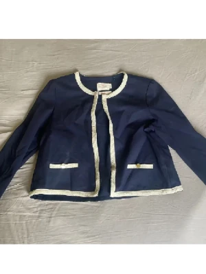 Mörkblå kavaj med vita detaljer - Snygg mörkblå kavaj från Holly&white med vita kantdetaljer och dekorativa knappar på fickorna. Jackan har rund halsringning, lång ärm och är tillverkad i bomull. Perfekt för dig som gillar klassisk stil med en twist. Passar s/m