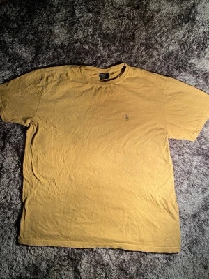 Beige t-shirt från Polo Ralph Lauren - Säljer en beige t-shirt från Polo Ralph Lauren i storlek L. Klassisk design med korta ärmar och rund hals. T-shirten har den ikoniska broderade Polo-loggan på bröstet och är gjord i mjuk bomull. Perfekt för dig som gillar stilrena och enkla plagg.