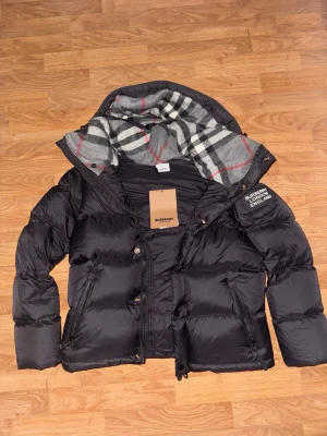 Svart pufferjacka från Burberry - Säljer en svart pufferjacka från Burberry med klassiskt rutigt foder i grått, vitt och rött. Jackan har dragkedja, knappar och två sidofickor med dragkedja. Märkeslogga på ärmen och varm, vadderad design. Passar alla 175cm och neråt 