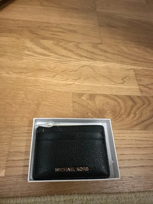 Svart korthållare från Michael Kors - Säljer en stilren svart korthållare från Michael Kors i äkta skinn. Korthållaren har flera kortfack och en minimalistisk design med guldfärgad logotyp framtill. Perfekt för dig som vill ha koll på korten och samtidigt hålla det classy.