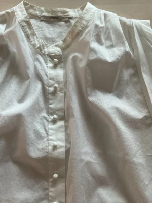 Vit henley-skjorta från Manoukian - Säljer en vit henley-skjorta från Manoukian i tunn bomull. Skjortan har rundad hals, dekorativa tygklädda knappar framtill och längre, avslappnad passform. Perfekt till lager på lager och enkel att styla med olika accessoarer.