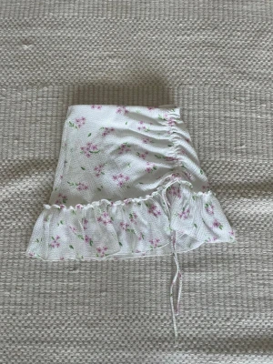 Vit blommig minikjol från Gina Tricot - Helt ny supersöt vit minikjol från Gina Tricot med rosa blommor och gröna blad. Kjolen har volangkant nedtill och snörning på sidan som ger en snygg rynkad effekt. Materialet är strukturerad polyester som känns lätt och fräsch. Perfekt för sommarens alla dagar!