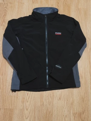 Svart vindjacka Peak Performance - Svart och grå vindjacka från Peak Performance, modell R&D. Jackan har hel dragkedja framtill, högt krage och två sidofickor med dragkedja. Materialet är syntet och jackan har Windstopper-detalj på ärmen. Perfekt för utomhusaktiviteter under kyliga dagar.