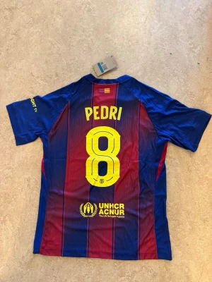 Fc Barcelona 25/26 Henmatröja  - FC Barcelona matchtröja 25/26  med Pedri och nummer 8 på ryggen, tillverkad av Nike. Tröjan har rödblåa ränder, gula detaljer och sponsorer som Spotify och UNHCR. 