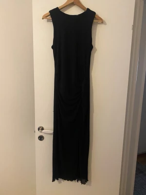 Svart långklänning ärmlös - Snygg svart långklänning med ärmlös design och rund halsringning. Klänningen har en rak siluett och lätt rynkade detaljer längs sidorna. Perfekt för dig som gillar stilrena och enkla plagg med en elegant touch. Den har även slits 