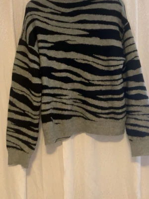 Grå och svart stickad zebramönstrad tröja - Säljer en stickad tröja med zebramönster i grått och svart. Tröjan har långärmade och ribbade muddar vid ärmslut och nederkant. Perfekt för dig som gillar djurmönster och vill ha en cool look.