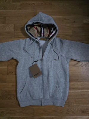 Grå burberry zip hoodie - En grå burberry zip hoodie i storlek M. Helt ny. Den är lite liten i storleken då jag är 175 och den sitter perfekt så hade varit bra för dig som är 170