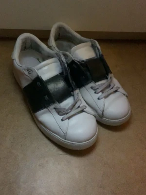 Valentino sneakers med svart band - Säljer ett par vita Valentino sneakers med ett brett svart band över mitten och coola nitar på hälen. Skorna har snörning och rund tå, tillverkade i skinn för en lyxig känsla. Perfekt för dig som gillar stilrena och exklusiva sneakers. och man kan få tjejer med de kanske lätt