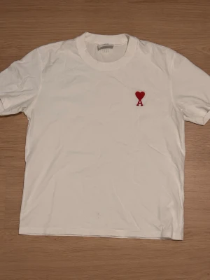 Vit t-shirt från Ami Paris med rött hjärta - Vit t-shirt från Ami paris med klassisk rund hals och korta ärmar. På bröstet finns ett broderat rött hjärta med bokstaven A. Tillverkad i mjuk bomull, perfekt för en clean och stilren look.
