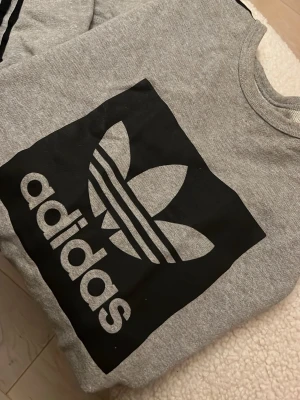 Grå sweatshirt från Adidas - Säljer en grå sweatshirt från Adidas med stor svart logga framtill. Tröjan har rund hals och är tillverkad i mjuk bomullsmix. Perfekt för en avslappnad och sportig stil. Passar dig som gillar klassiska streetwear-plagg.