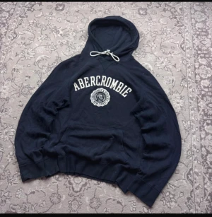 Blå hoodie Abercrombie & Fitch M - Mörkblå hoodie från Abercrombie & Fitch i storlek M. Tröjan har vit broderad logga och emblem på bröstet, huva med vit snörning och en klassisk känguruficka framtill. Perfekt för chill dagar och streetwear-stil.